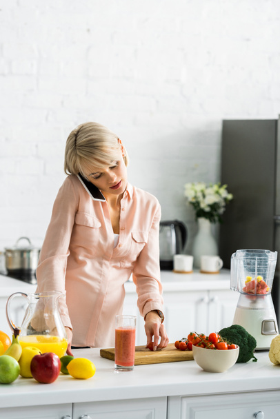 blonde pregnant woman talking on smartphone in kitchen near cherry tomatoes on cutting board - Photo, Image