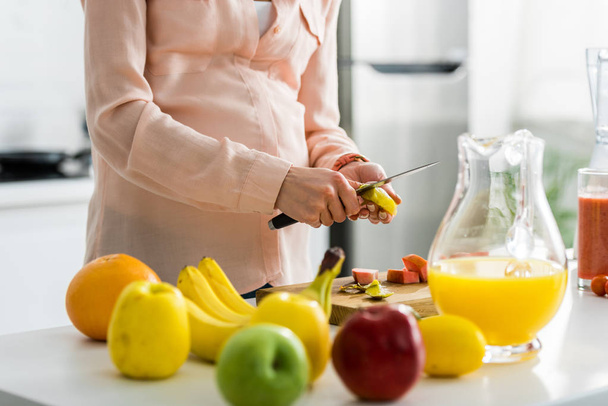 обрезанный вид беременной молодой женщины, обрезающей фрукты на кухне
  - Фото, изображение