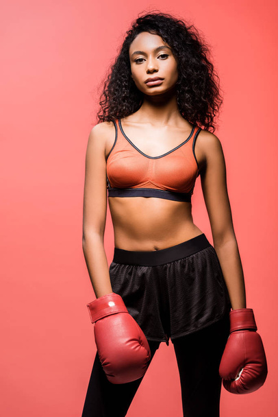 beautiful african american sportswoman in boxing gloves looking at camera and posing isolated on coral - Valokuva, kuva