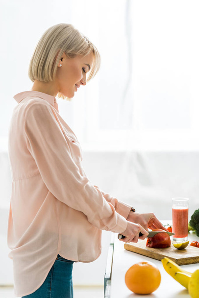 happy blonde pregnant woman cutting apple on chopping board  - Photo, Image