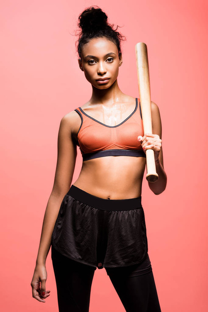 beautiful african american sportswoman looking at camera and posing with baseball bat isolated on coral  - Фото, зображення
