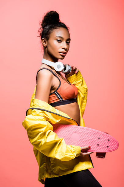 beautiful african american sportswoman with headphones looking at camera while holding penny board isolated on coral - Fotoğraf, Görsel
