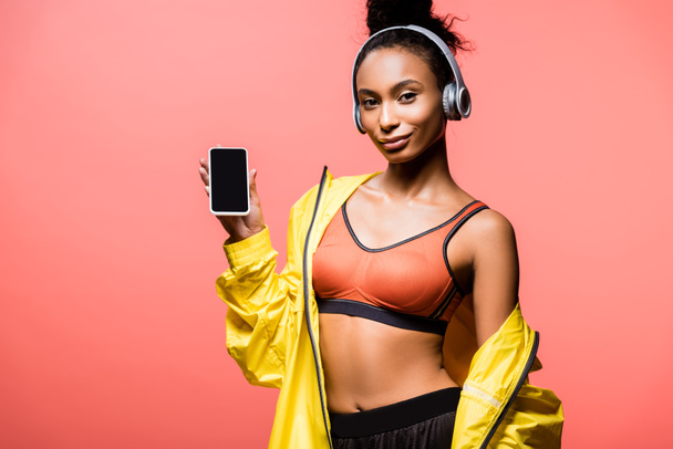krásná africká americká sportovkyně ve sluchátkách při pohledu na fotoaparát a prezentaci smartphone s prázdnou obrazovkou izolované na coral - Fotografie, Obrázek