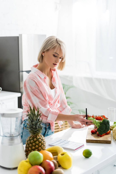 blondes Mädchen hält Stift und Bio-Brokkoli in der Nähe von Zutaten in der Küche  - Foto, Bild