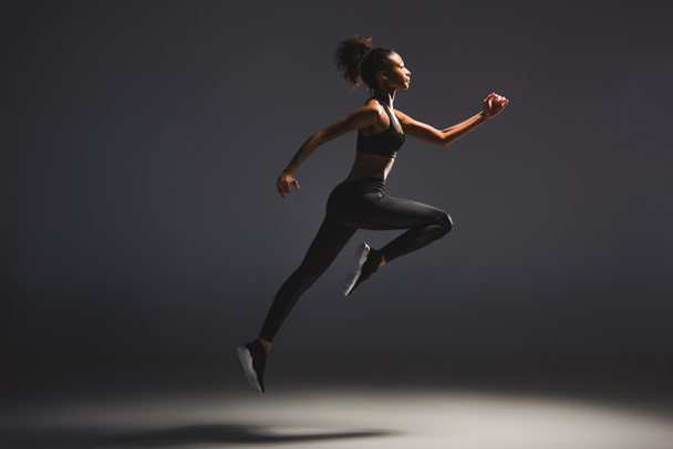 side view of beautiful athletic african american sportswoman jumping on black - Photo, Image