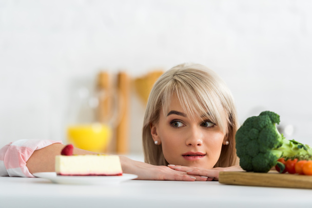 blonde girl looking at saucer with sweet cake near organic vegetables  - Fotoğraf, Görsel