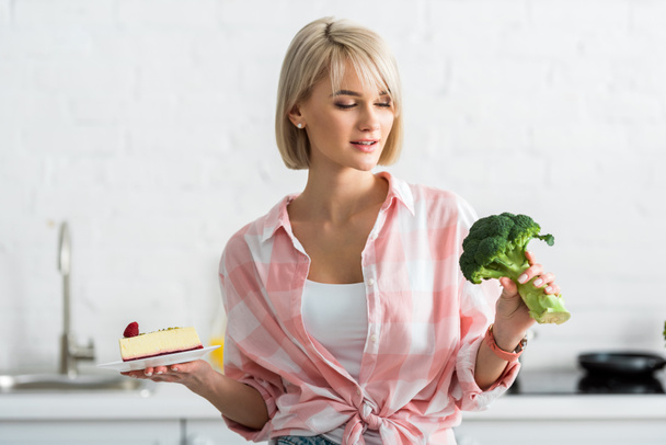 blonde girl looking at organic broccoli while holding saucer with sweet cake - Foto, imagen