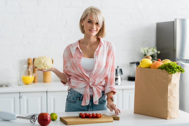 cheerful blonde girl holding cauliflower near paper bag with groceries - 写真・画像
