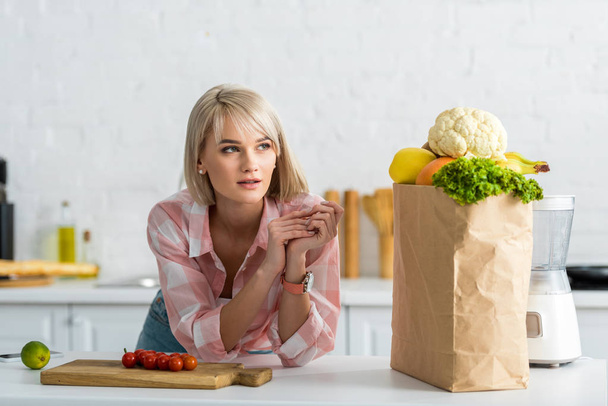 dreamy blonde woman near paper bag with groceries in kitchen  - Foto, imagen