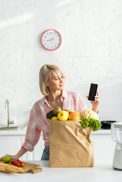 happy blonde woman holding smartphone with blank screen near paper bag with ingredients   - Photo, Image