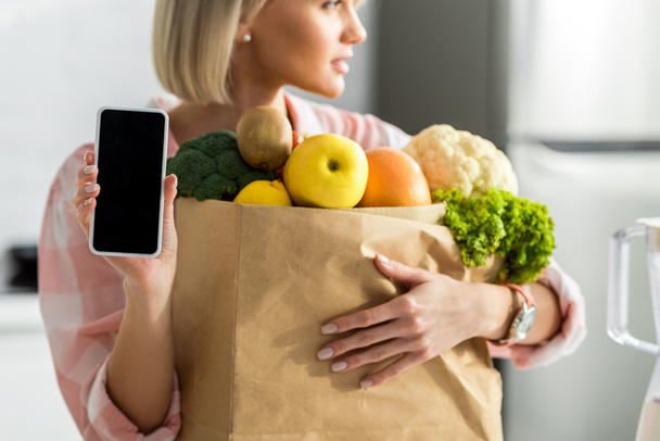 cropped view of blonde girl holding smartphone with blank screen near paper bag with groceries    - Photo, Image