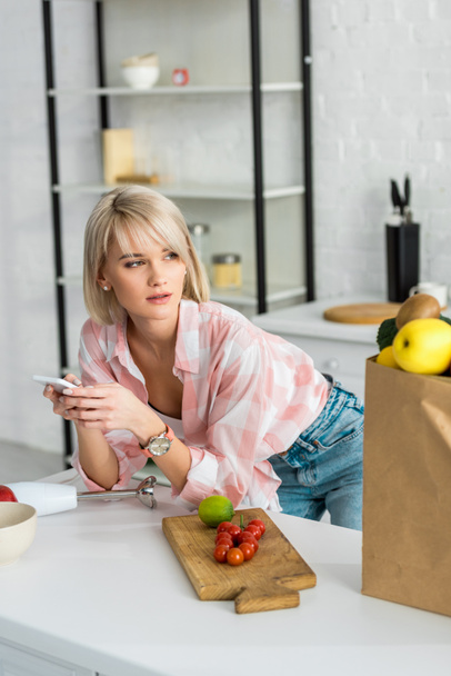 Blondes Mädchen benutzt Smartphone in der Nähe von Papiertüte mit Lebensmitteln    - Foto, Bild