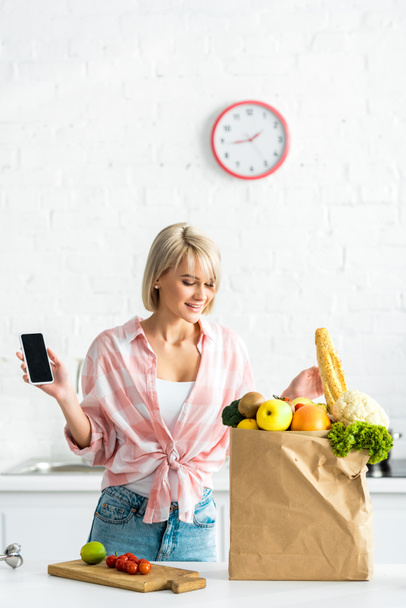 boldog lány gazdaság okostelefon üres képernyő közelében papírzacskót az Élelmiszeri    - Fotó, kép