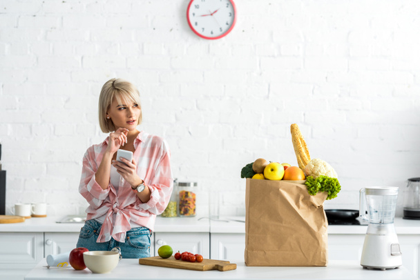 pensive blonde girl with smartphone near paper bag with groceries  - Fotó, kép