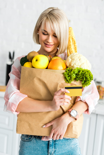 happy blonde girl holding credit card while hugging paper bag with groceries  - Fotoğraf, Görsel
