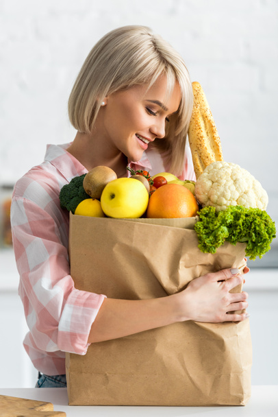 χαρούμενη ξανθιά γυναίκα που αγκαλιάζει χάρτινη σακούλα με λαχανικά  - Φωτογραφία, εικόνα