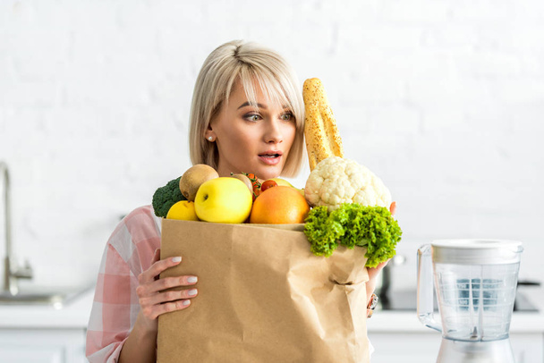 mujer joven rubia sorprendida abrazando bolsa de papel con comestibles y mirando licuadora
  - Foto, imagen