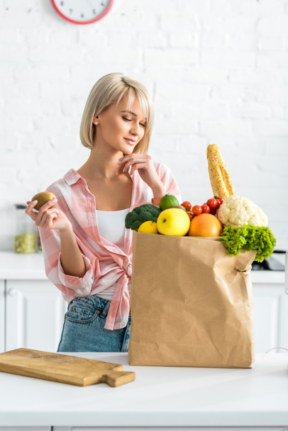 happy blonde young woman holding kiwi fruit near paper bag with groceries - Fotó, kép