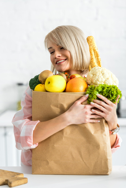 ευτυχισμένη ξανθιά νεαρή γυναίκα αγκαλιάζει χάρτινη σακούλα με λαχανικά - Φωτογραφία, εικόνα