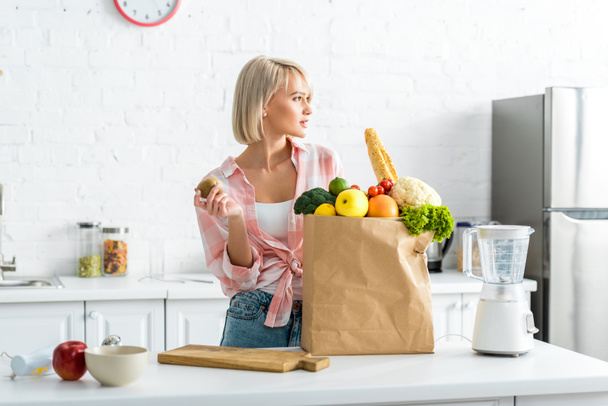 attraktive blonde Frau mit Kiwi-Früchten in der Nähe von Papiertüten mit Lebensmitteln  - Foto, Bild