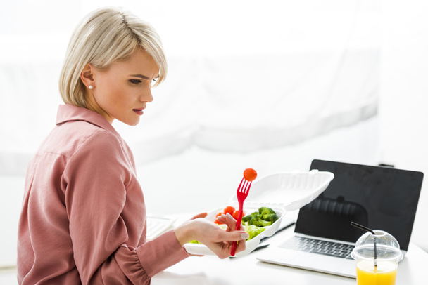 blonde freelancer near laptop with blank screen and vegetables in takeaway box - Photo, Image