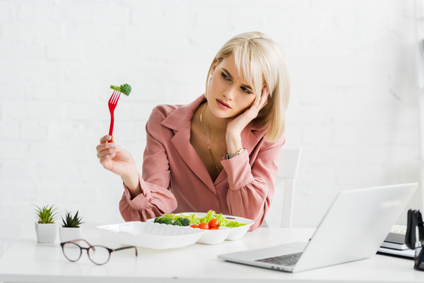 upset freelancer holding fork with cherry tomato near laptop - Foto, imagen