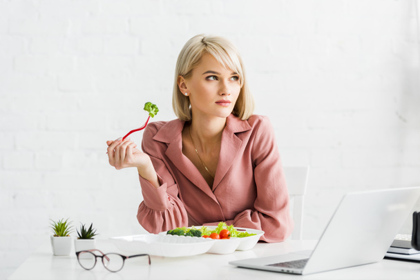 attractive freelancer holding fork with broccoli near laptop - Photo, Image