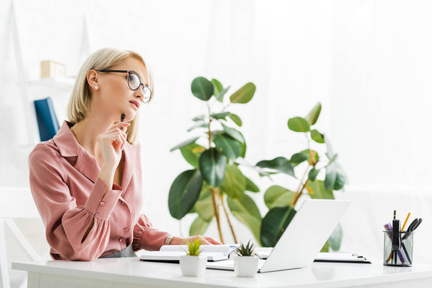 pensive young blonde freelancer in glasses sitting near laptop  - Фото, изображение