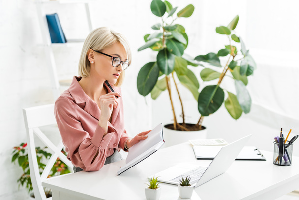 pensive young blonde freelancer in glasses holding pen and notebook while sitting near laptop  - Foto, Bild
