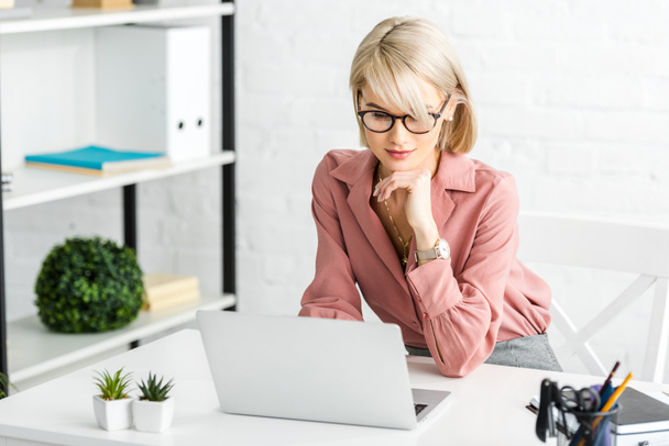 pensive young blonde freelancer in glasses looking at laptop  - Foto, Imagem