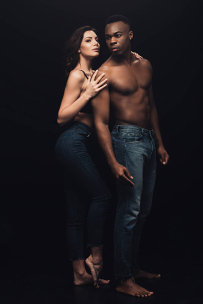 beautiful seductive woman hugging shirtless african american man in denim isolated on black - Foto, afbeelding