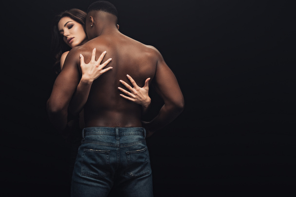 back view of beautiful sexy woman hugging shirtless african american man isolated on black - 写真・画像