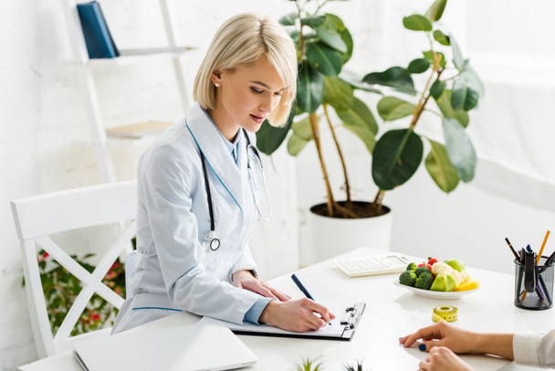 nutricionista loira atraente no diagnóstico de escrita casaco branco perto do paciente
  - Foto, Imagem