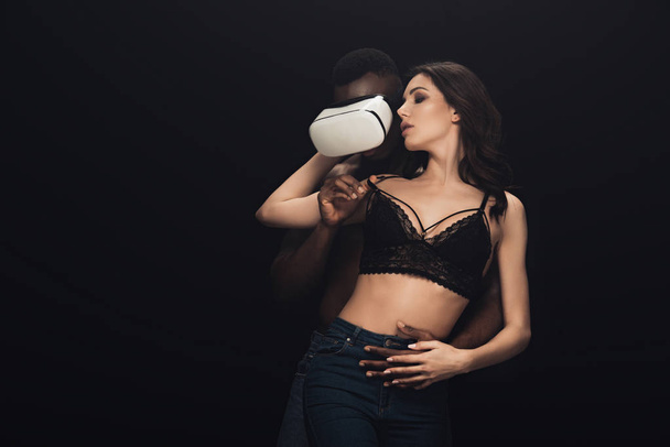 african american man in virtual reality headset hugging sexy woman isolated on black with copy space - Photo, Image