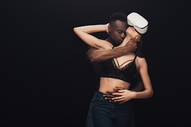 Afrikanische amerikanische Mann umarmt und küsst sexy Frau in Virtual-Reality-Headset isoliert auf schwarz - Foto, Bild