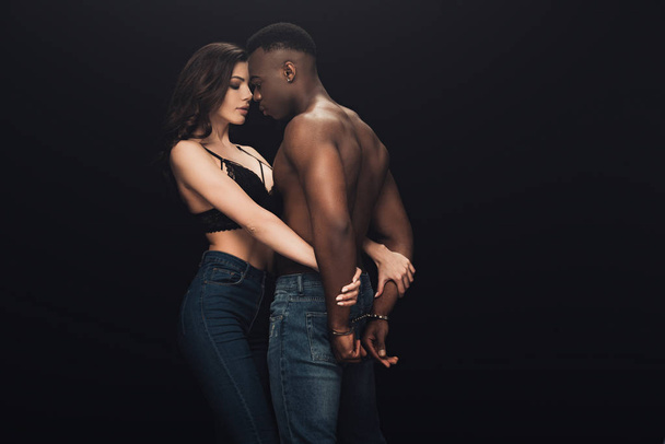 beautiful sexy woman hugging shirtless african american man in handcuffs isolated on black with copy space - Zdjęcie, obraz