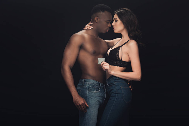 Krásná sexy žena, která objímá širý africký americký muž a drží kondom izolovaný na černém - Fotografie, Obrázek