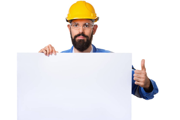Bauarbeiter im Bauhelm hebt den Daumen und hält eine weiße Werbetafel mit Kopierraum in die Höhe, isoliert - Foto, Bild