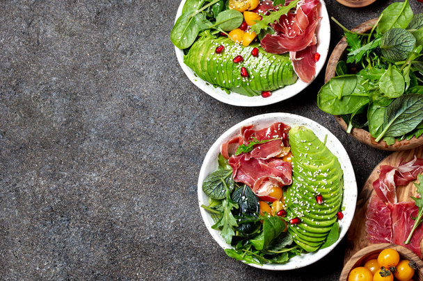 Low carbs bowl. Fresh salad with green spinach, rucola, avocado an ham serrano in white bowl, gray background, top view - 写真・画像
