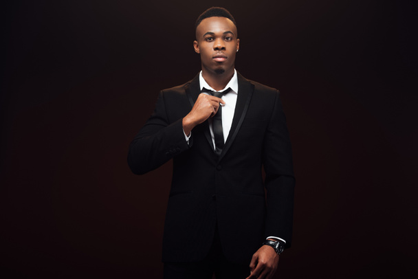 handsome african american man in suit adjusting tie and looking at camera isolated on black  - Photo, Image