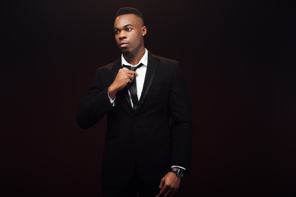 handsome fashionable african american man in suit adjusting tie isolated on black  - Zdjęcie, obraz