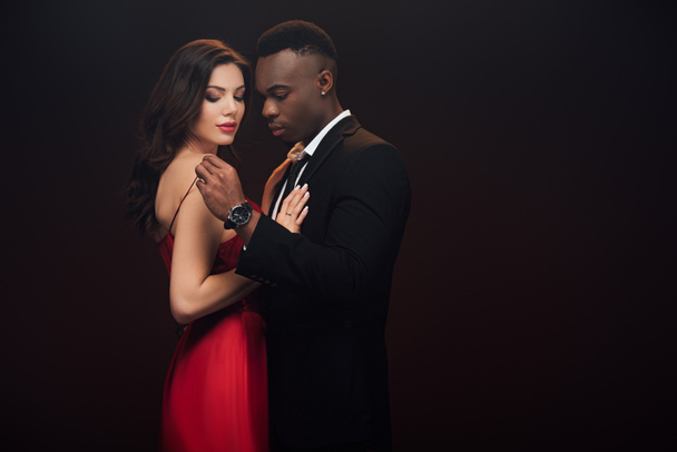 beautiful sensual interracial couple in formal wear embracing isolated on black with copy space - Фото, изображение