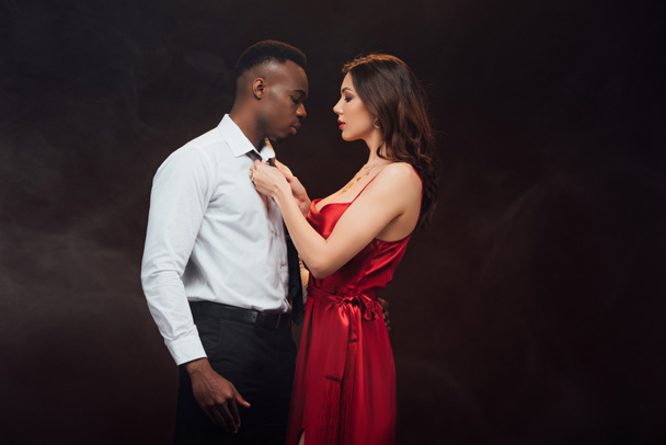 beautiful woman in red dress undressing shirt of african american man isolated on black with copy space - Fotoğraf, Görsel