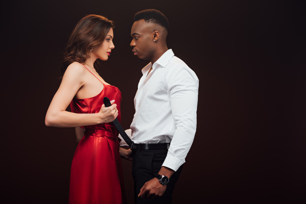 attractive woman in red dress undressing african american man isolated on black with copy space - Photo, Image