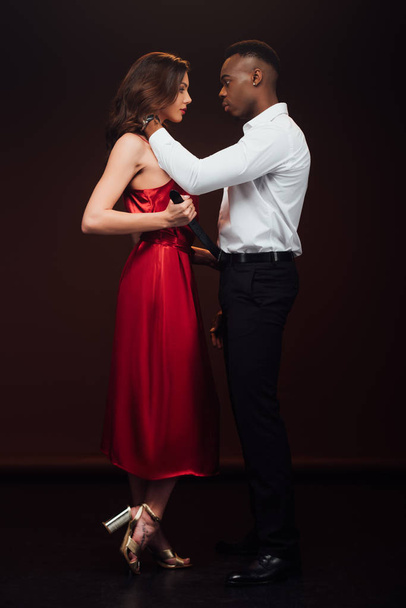 handsome african american man touching neck of beautiful woman in red dress isolated on black - Zdjęcie, obraz