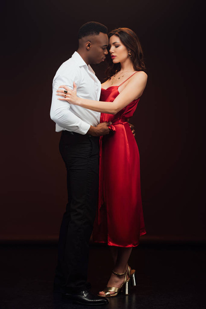 handsome african american man embracing beautiful woman in red dress isolated on black - Zdjęcie, obraz