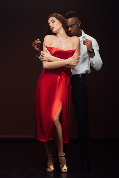 african american man undressing beautiful woman in red dress isolated on black - Photo, Image