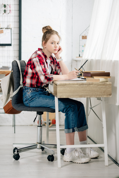Pensive teenager in checkered shirt holding pen at table while doing homework - Foto, Bild