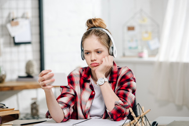 Sad teenager in checkered shirt and headphones looking at smartphone during video call - Фото, изображение