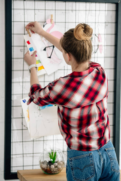 Back view of teenager in checkered shirt hanging drawing on pinboard - Foto, afbeelding